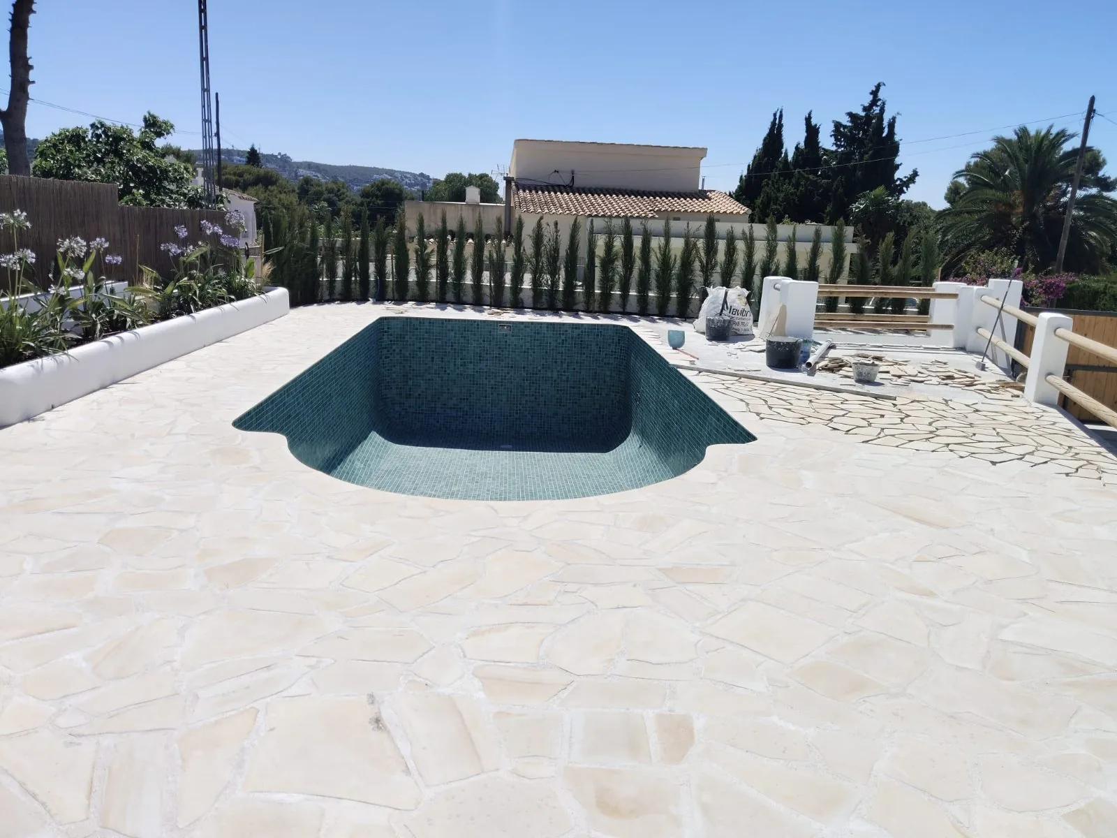 Piscina con mosaico verde rodeada de amplia terraza de piedra natural y cipreses