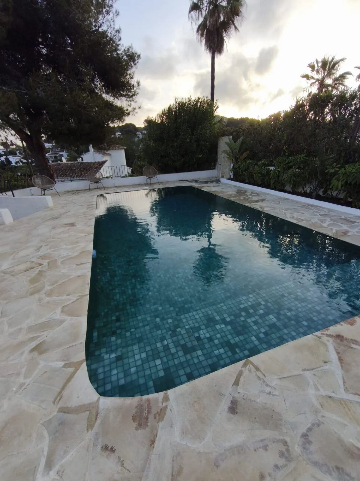 Piscina de mosaico verde oscuro con terraza de piedra clara al atardecer