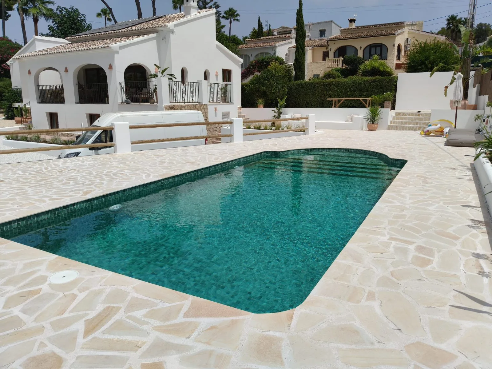Piscina de mosaico verde con terraza de piedra clara y villa blanca al fondo